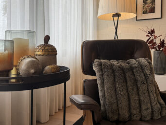 Gemütlich beleuchtetes Wohnzimmer im Herbst-Look mit warmem Kerzenlicht, Laterne, Kürbis und herbstlicher Deko auf einem Holztisch. Im Hintergrund sorgt eine Tischlampe für indirektes Licht und schafft eine einladende, stimmungsvolle Atmosphäre – perfekt für Interior- und Wohninspiration im Herbst und Winter.