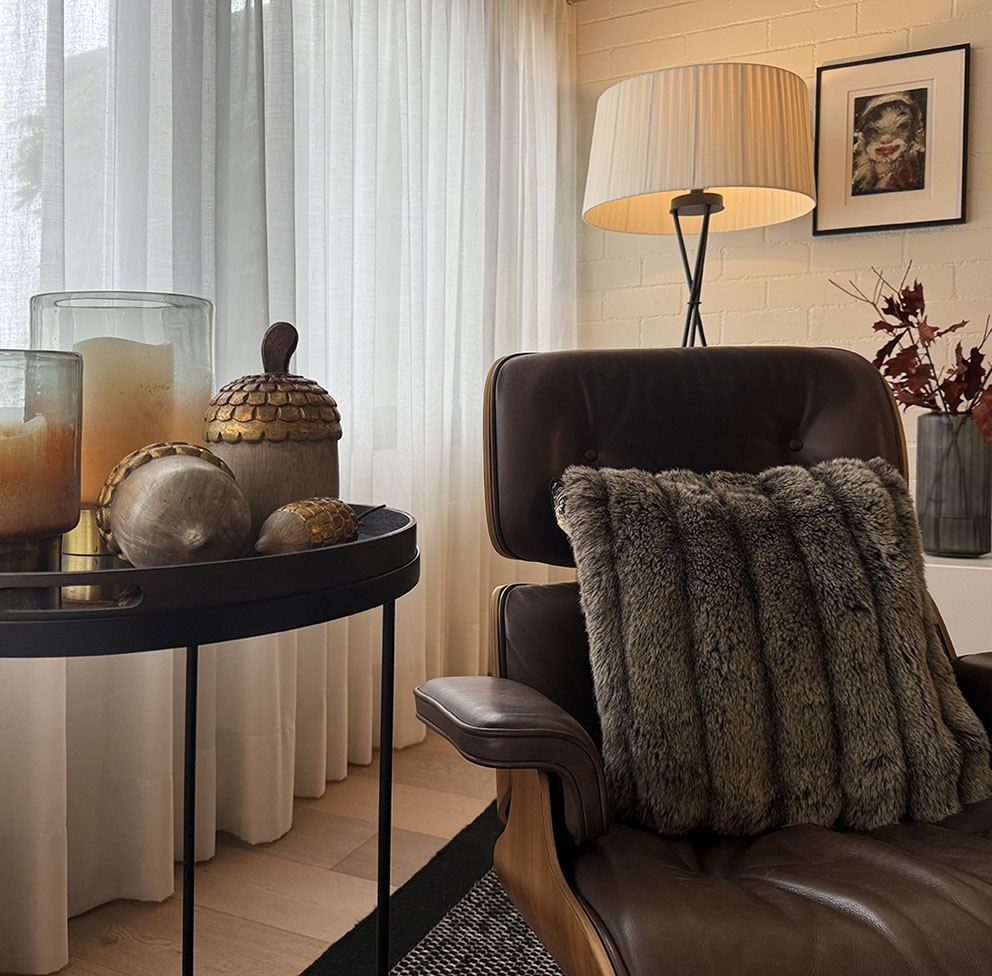 Gemütlich beleuchtetes Wohnzimmer im Herbst-Look mit warmem Kerzenlicht, Laterne, Kürbis und herbstlicher Deko auf einem Holztisch. Im Hintergrund sorgt eine Tischlampe für indirektes Licht und schafft eine einladende, stimmungsvolle Atmosphäre – perfekt für Interior- und Wohninspiration im Herbst und Winter.