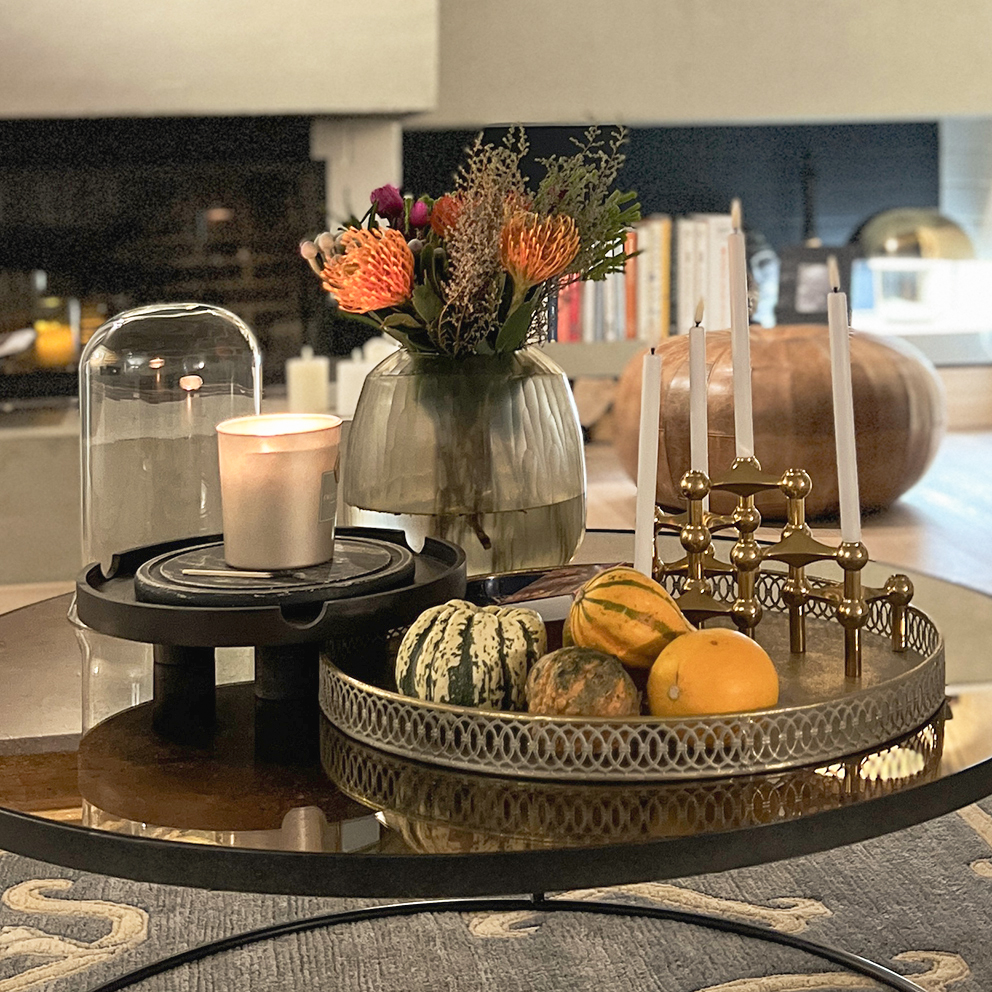 Gemütlich beleuchtetes Wohnzimmer im Herbst-Look mit warmem Kerzenlicht, Laterne, Kürbis und herbstlicher Deko auf einem Holztisch. Im Hintergrund sorgt eine Tischlampe für indirektes Licht und schafft eine einladende, stimmungsvolle Atmosphäre – perfekt für Interior- und Wohninspiration im Herbst und Winter.